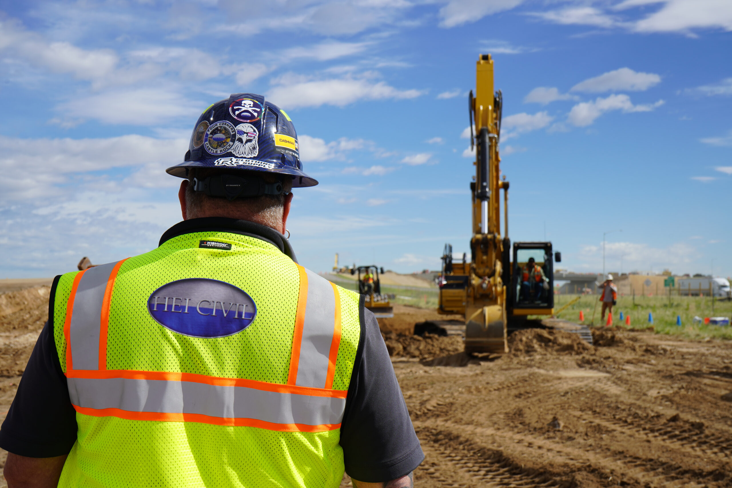 An HEI Civil team member looking on at a job site while an excavator digs.