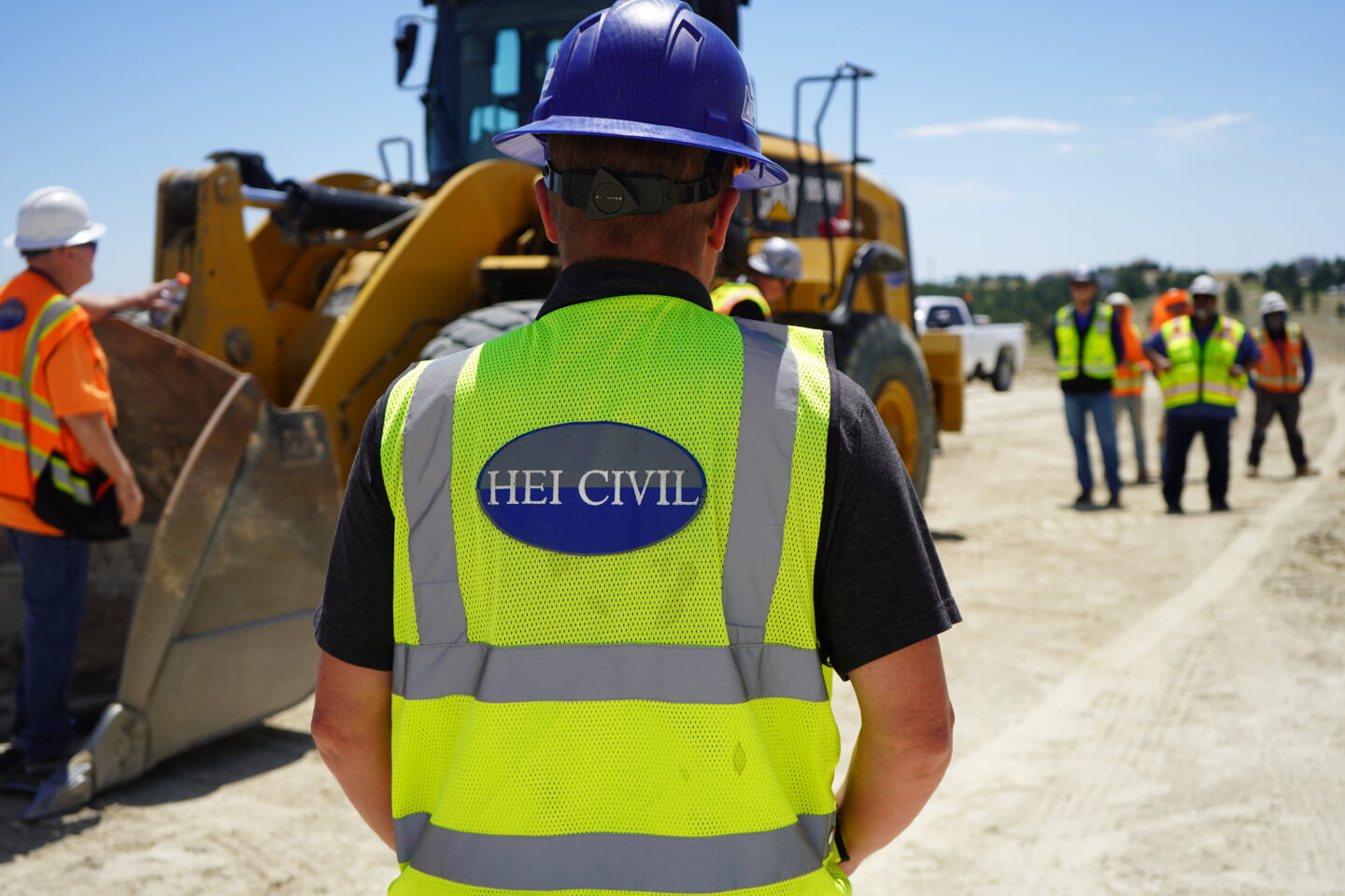 Employee wearing a high visible vest and hard hat, pictured from behind. Getting hands-on training in the field, next to a wheel loader.
